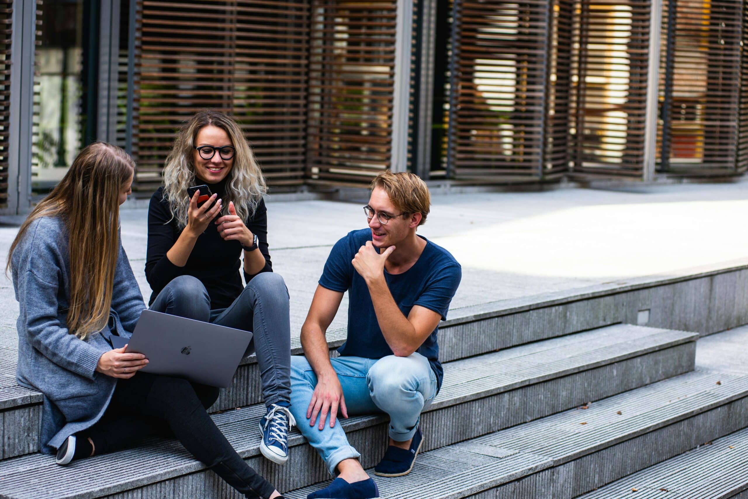 Drei junge Studierende sitzen entspannt auf Treppenstufen, lachen gemeinsam und nutzen Smartphone und Laptop – moderne, studentische Atmosphäre in Hannover